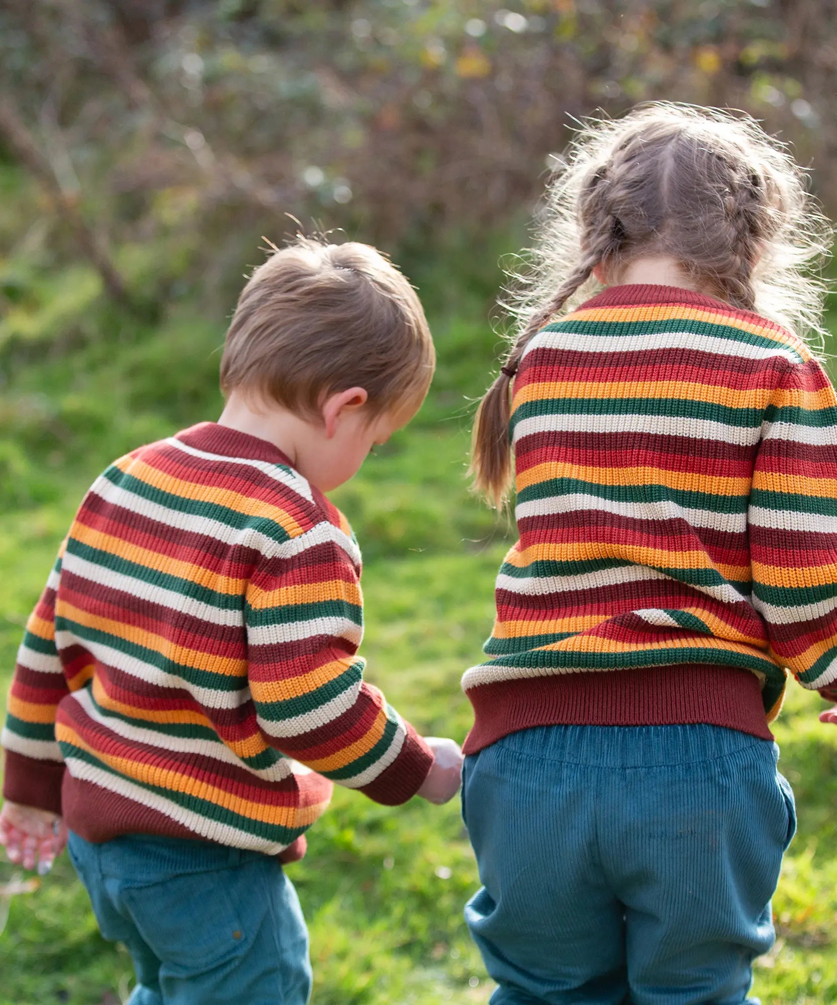 Two children wearing the Little Green Radicals knitted snuggly jumper in a  hazel rainbow striped design  with blue corduroy trousers. 