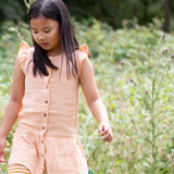 Child wearing Little Green Radicals coral pink dropped waist children's dress with ochre yellow pom pom ribbon edging on the sleeves and collar with striped leggings underneath.