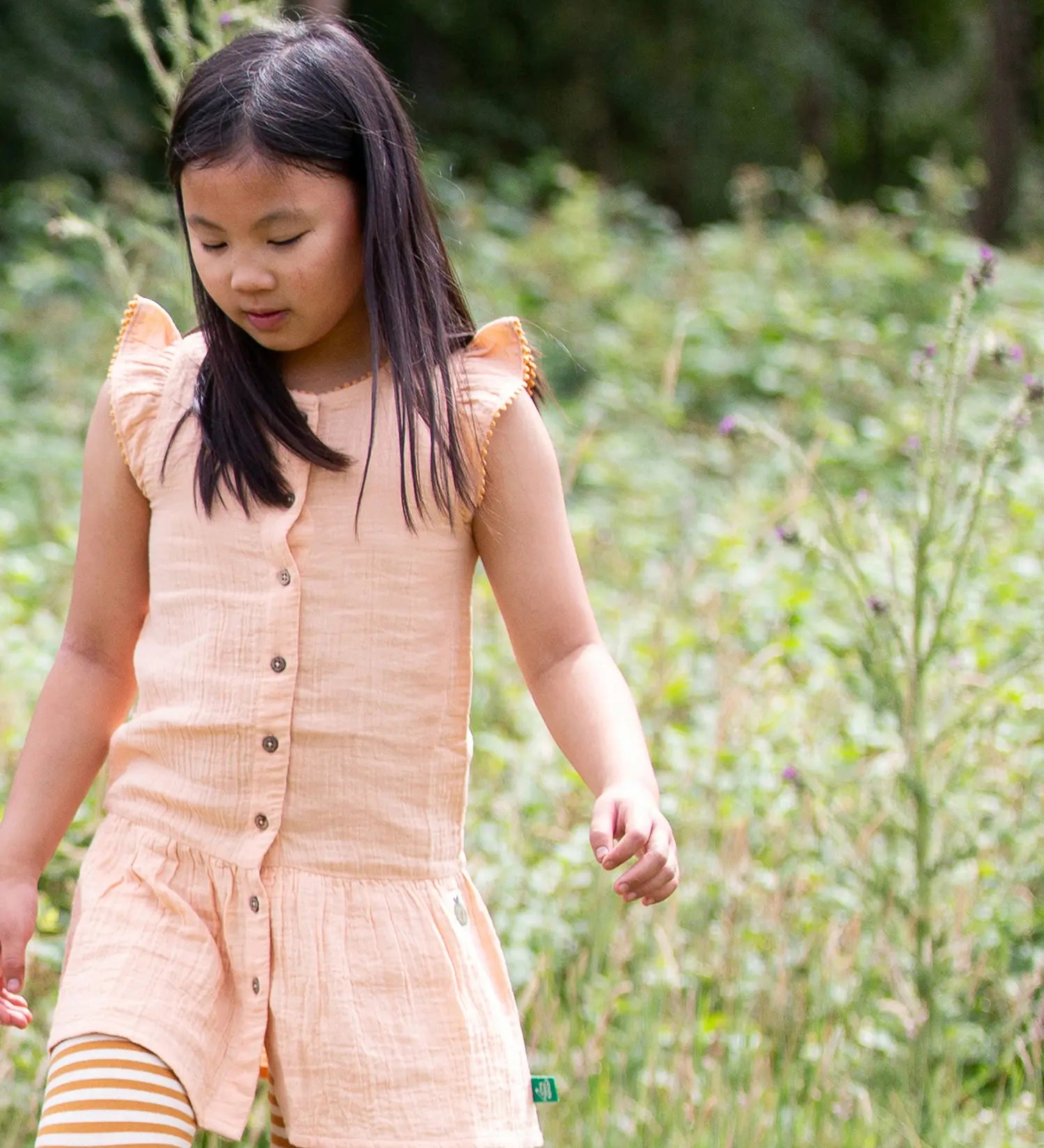Child wearing Little Green Radicals coral pink dropped waist children's dress with ochre yellow pom pom ribbon edging on the sleeves and collar with striped leggings underneath.