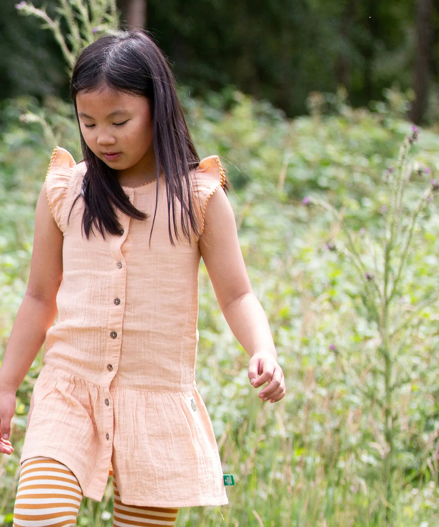 Child wearing Little Green Radicals coral pink dropped waist children's dress with ochre yellow pom pom ribbon edging on the sleeves and collar with striped leggings underneath.