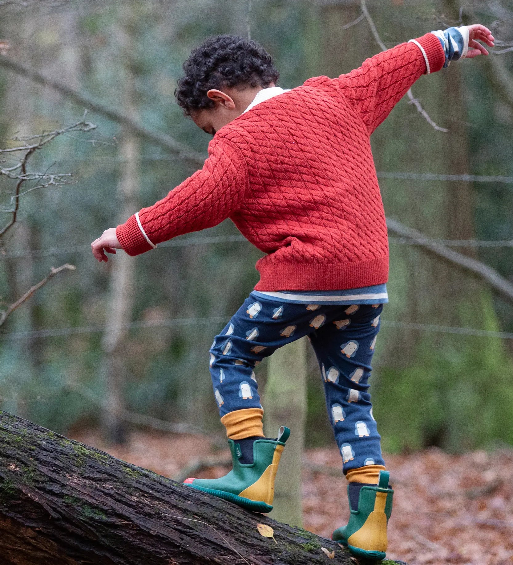 Child wearing Little Green Radicals organic cotton comfy joggers with a blue penguins design and a red knitted cardigan.