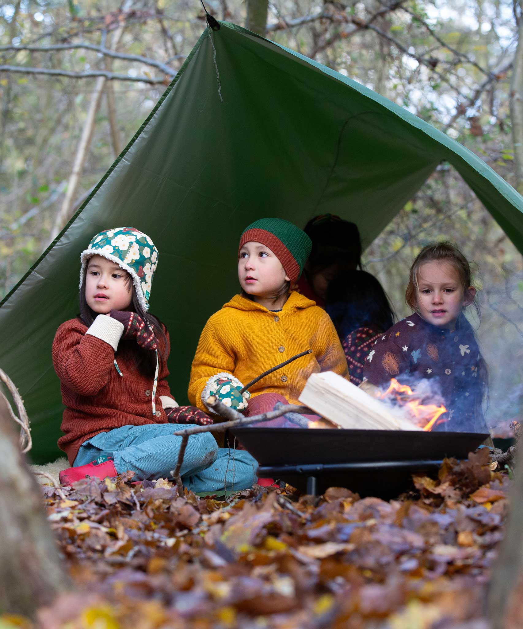 Three children sitting by a campfire wearing Little green Radicals knitwear and hats. These organic cotton clothing and accessories are part of a wide range available here at Babipur. 