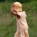 Back of a child wearing the Little Green Radicals coral pink dropped waist children's dress with ochre yellow pom pom ribbon edging on the sleeves and collar with a yellow gingham sun hat.
