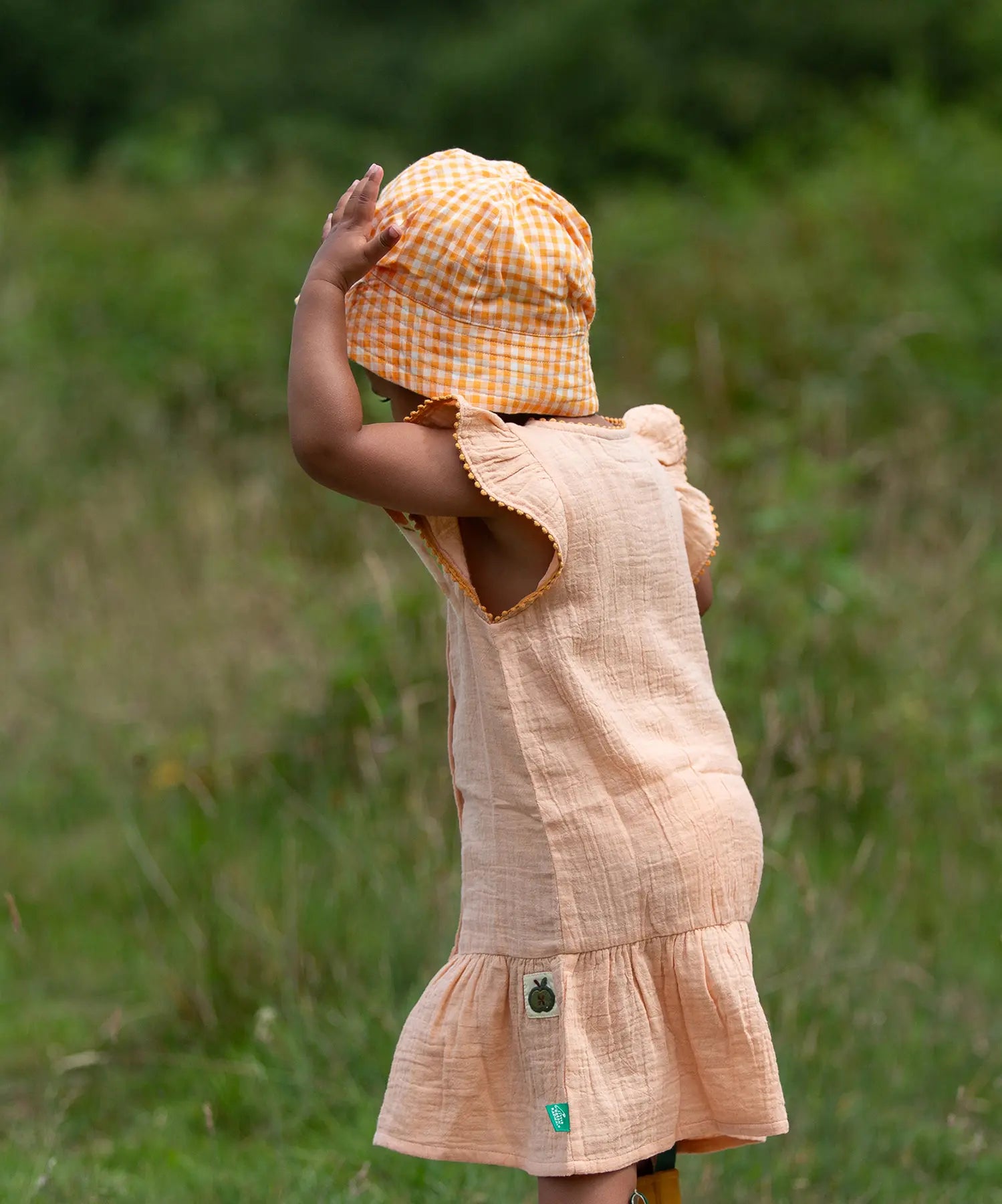 Back of a child wearing the Little Green Radicals coral pink dropped waist children's dress with ochre yellow pom pom ribbon edging on the sleeves and collar with a yellow gingham sun hat.