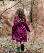 Back of a child wearing the Little Green Radicals pink flowers sailor dress in purple with green wellington boots. 