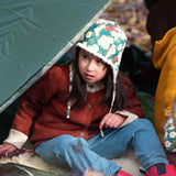 A child sitting by a campfir wearing a Little Green Radicals Children's Hedgerow print Sherpa Fleece Cosy Hat with a ginger coloured knitted cardigan. These lined hats are part  of a wide range of organic cotton clothing for children available here at Babipur. 