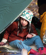 A child sitting by a campfir wearing a Little Green Radicals Children's Hedgerow print Sherpa Fleece Cosy Hat with a ginger coloured knitted cardigan. These lined hats are part  of a wide range of organic cotton clothing for children available here at Babipur. 