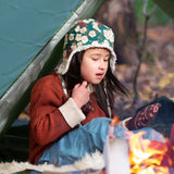 A child sitting by a campfir wearing a Little Green Radicals Children's Hedgerow print Sherpa Fleece Cosy Hat with a ginger coloured knitted cardigan. These lined hats are part  of a wide range of organic cotton clothing for children available here at Babipur. 