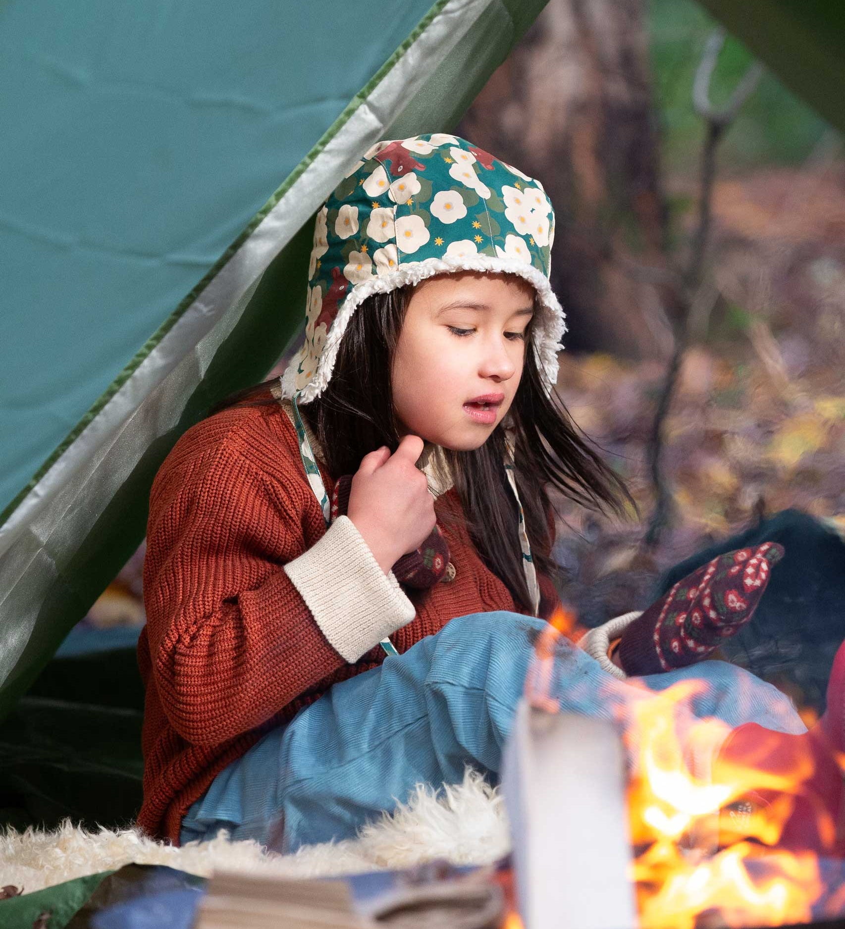 A child sitting by a campfir wearing a Little Green Radicals Children's Hedgerow print Sherpa Fleece Cosy Hat with a ginger coloured knitted cardigan. These lined hats are part  of a wide range of organic cotton clothing for children available here at Babipur. 