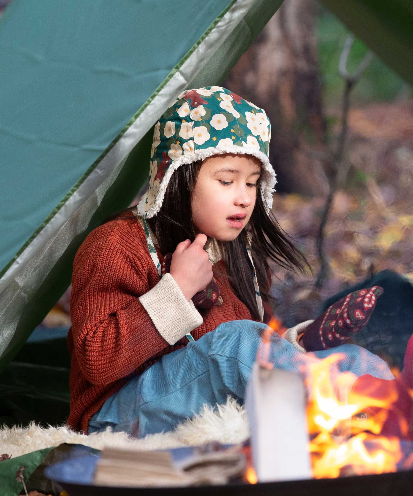 A child sitting by a campfir wearing a Little Green Radicals Children's Hedgerow print Sherpa Fleece Cosy Hat with a ginger coloured knitted cardigan. These lined hats are part  of a wide range of organic cotton clothing for children available here at Babipur. 