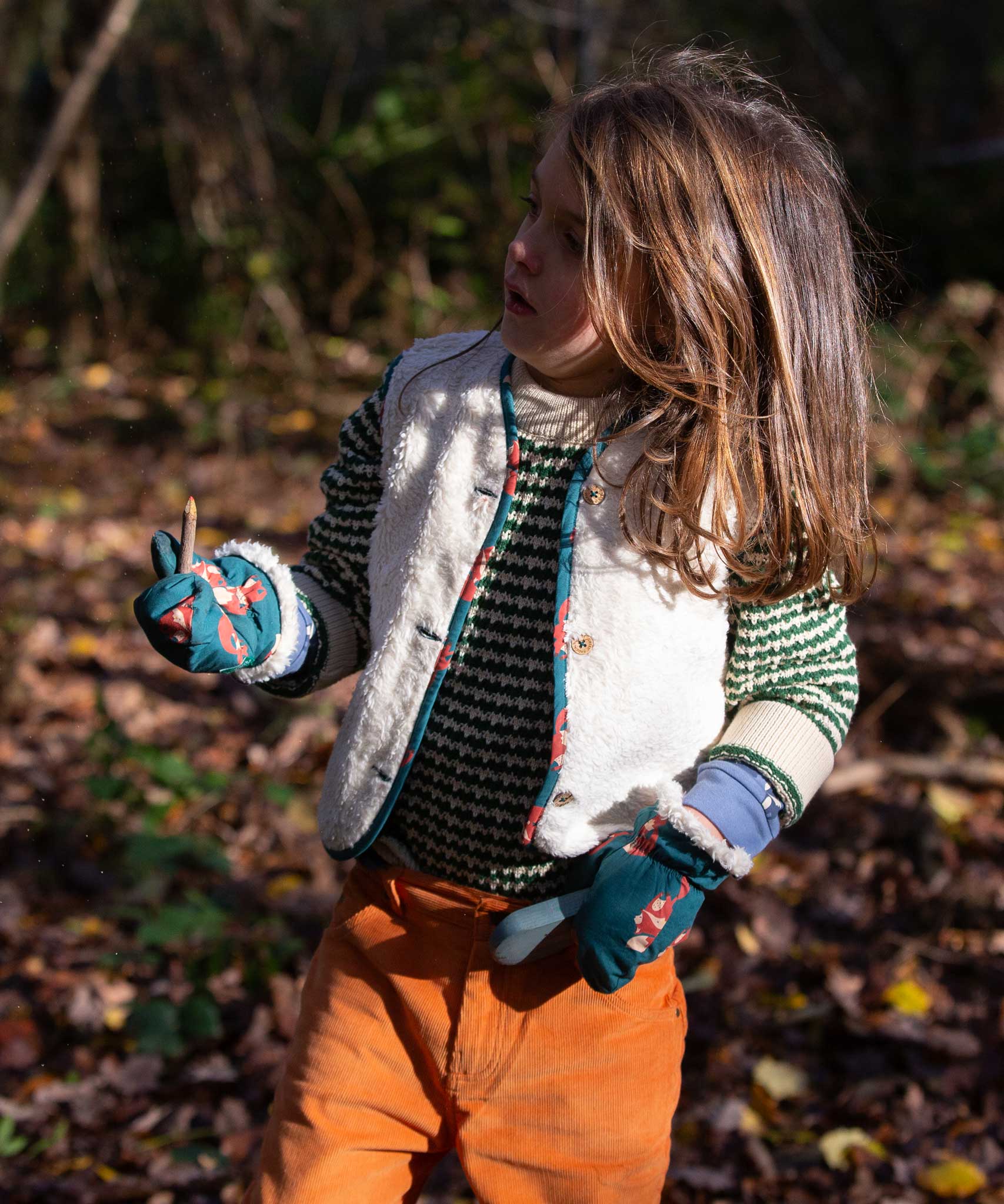 A child wearing the Little Green Radicals fox print sherpa fleece lined mittens with a knitted jumper and sherpa lined reversible gilet and yellow corduroy trousers. The children's sized mittens are part of a wide range of organic cotton clothing for children available here at Babipur. 
