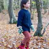 A child walking through a forest on a floor covered with fallen leaves wearing the  Little Green Radicals Hedgerow print Children's Sherpa Lined Mittens with a navy knitted jumper and ginger corduroy trousers.