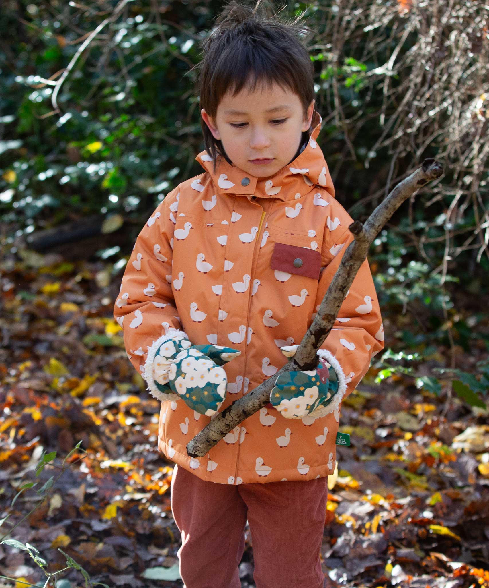 A child holding a stick wearing the  Little Green Radicals Hedgerow print Children's Sherpa Lined Mittens and a duck print coat. 