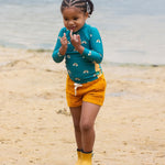 A child wearing the Little Green Radicals Teal Rainbows UPF 50+ Sunsafe Long Sleeve Rash Vest with yellow gold shorts and yellow wellingtons. The child is standing on a sandy beach. 
