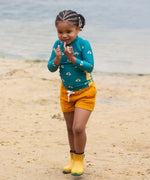 A child wearing the Little Green Radicals Teal Rainbows UPF 50+ Sunsafe Long Sleeve Rash Vest with yellow gold shorts and yellow wellingtons. The child is standing on a sandy beach. 