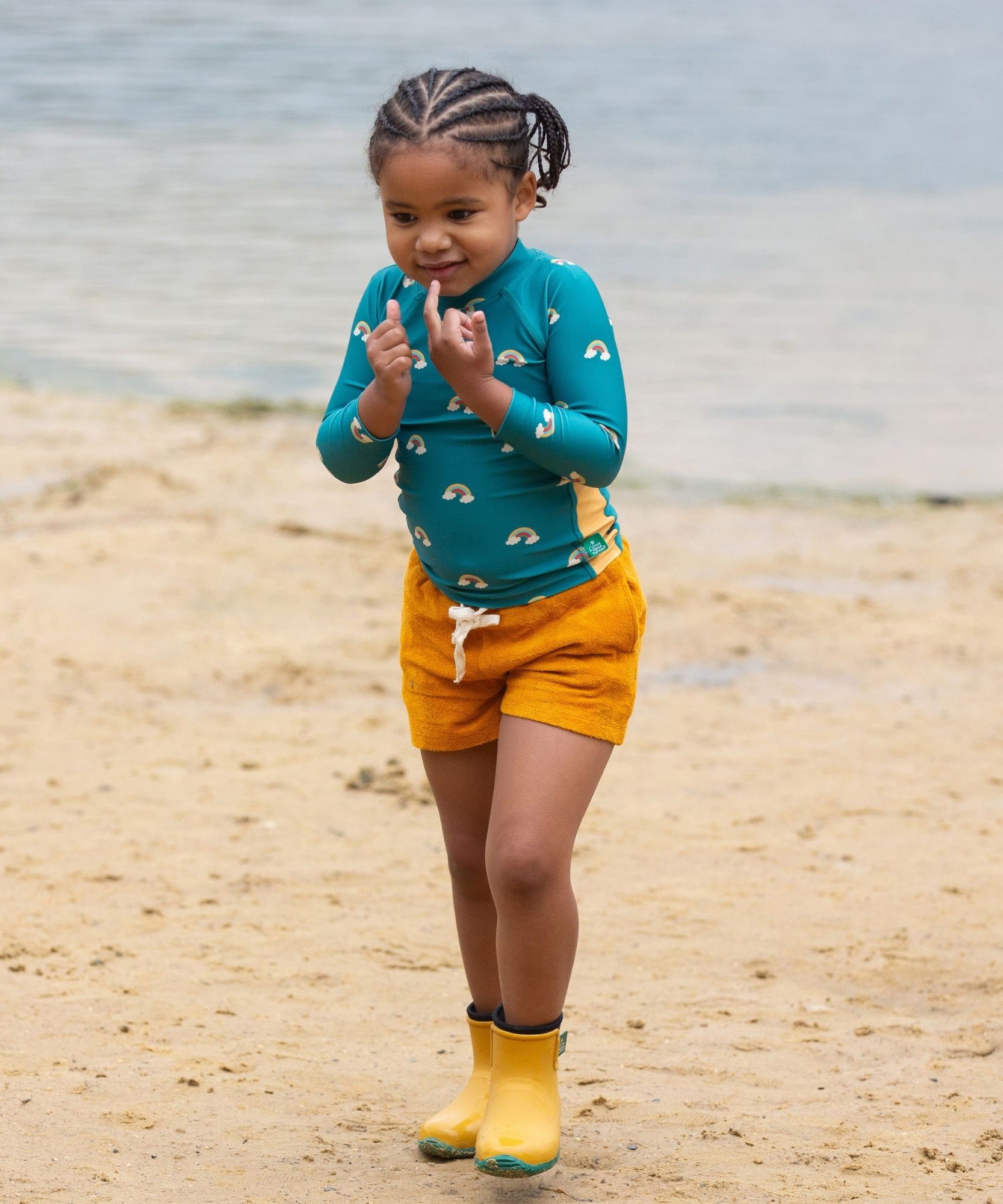 A child wearing the Little Green Radicals Teal Rainbows UPF 50+ Sunsafe Long Sleeve Rash Vest with yellow gold shorts and yellow wellingtons. The child is standing on a sandy beach. 