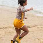 A child running along a sandy beach wearing the Little Green Radicals Vintage Flowers design UPF 50+ Sun safe Long Sleeve Rash Vest with yellow gold shorts and yellow wellingtons. 