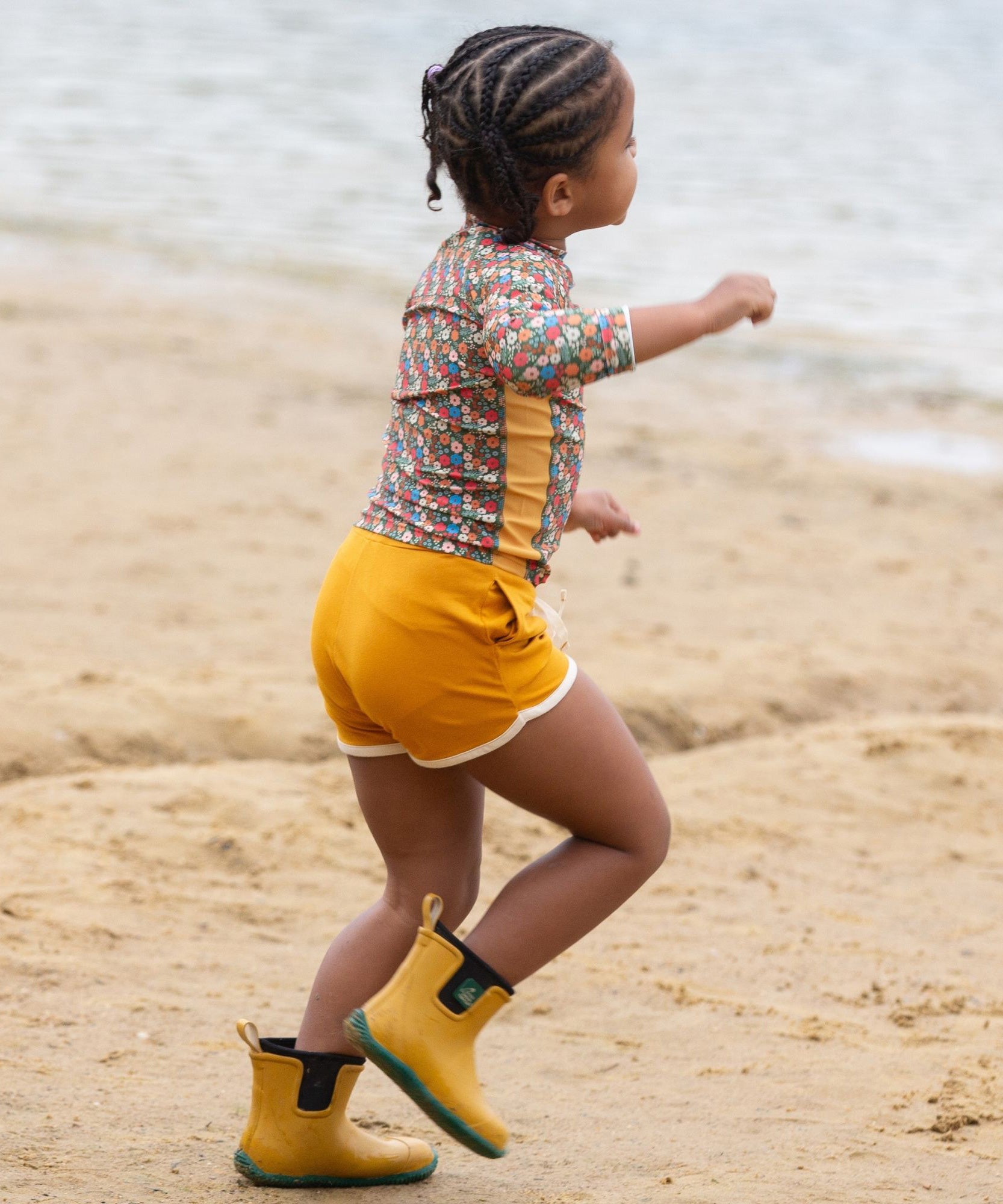 A child running along a sandy beach wearing the Little Green Radicals Vintage Flowers design UPF 50+ Sun safe Long Sleeve Rash Vest with yellow gold shorts and yellow wellingtons. 