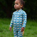 A child wearing the Little Green Radicals Blue Whales UPF 50+ Recycled Sunsafe Sunsuit with red wellington boots on grass outdoors.
