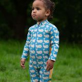A child wearing the Little Green Radicals Blue Whales UPF 50+ Recycled Sunsafe Sunsuit with red wellington boots on grass outdoors.