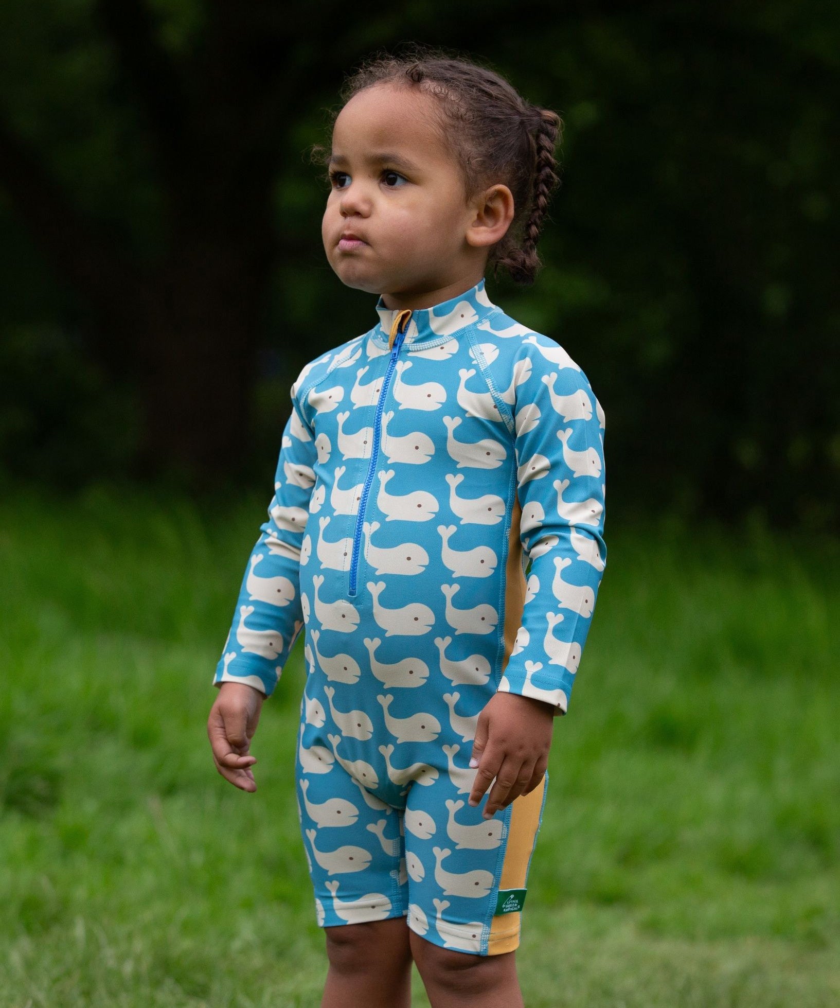 A child wearing the Little Green Radicals Blue Whales UPF 50+ Recycled Sunsafe Sunsuit with red wellington boots on grass outdoors.
