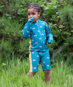 A child wearing the Little Green Radicals Teal Rainbows UPF 50+ Recycled Sunsafe Sunsuit standing in long grass.