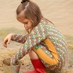 A child wearing the Little Green Radicals Vintage Flowers UPF 50+ Recycled Sunsafe Sunsuit with a pair of red wellington boots. The child is kneeling down on sand filling a bucket with sand. 