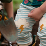 Close up of the Little Green Radicals weather knee patch blue and cream striped organic cotton joggers worn with a green apple print t-short and yellow wellington boots.