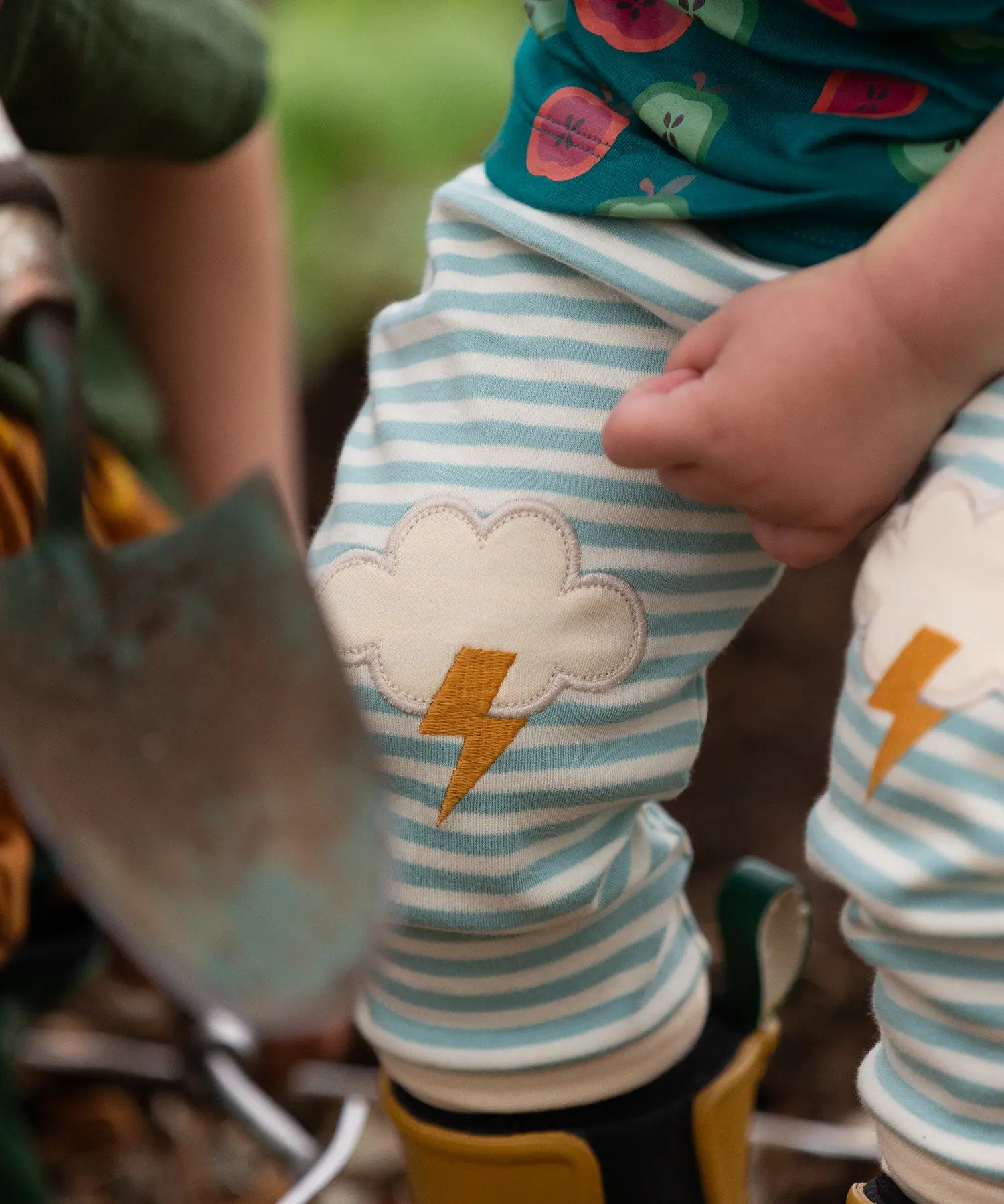 Close up of the Little Green Radicals weather knee patch blue and cream striped organic cotton joggers worn with a green apple print t-short and yellow wellington boots.