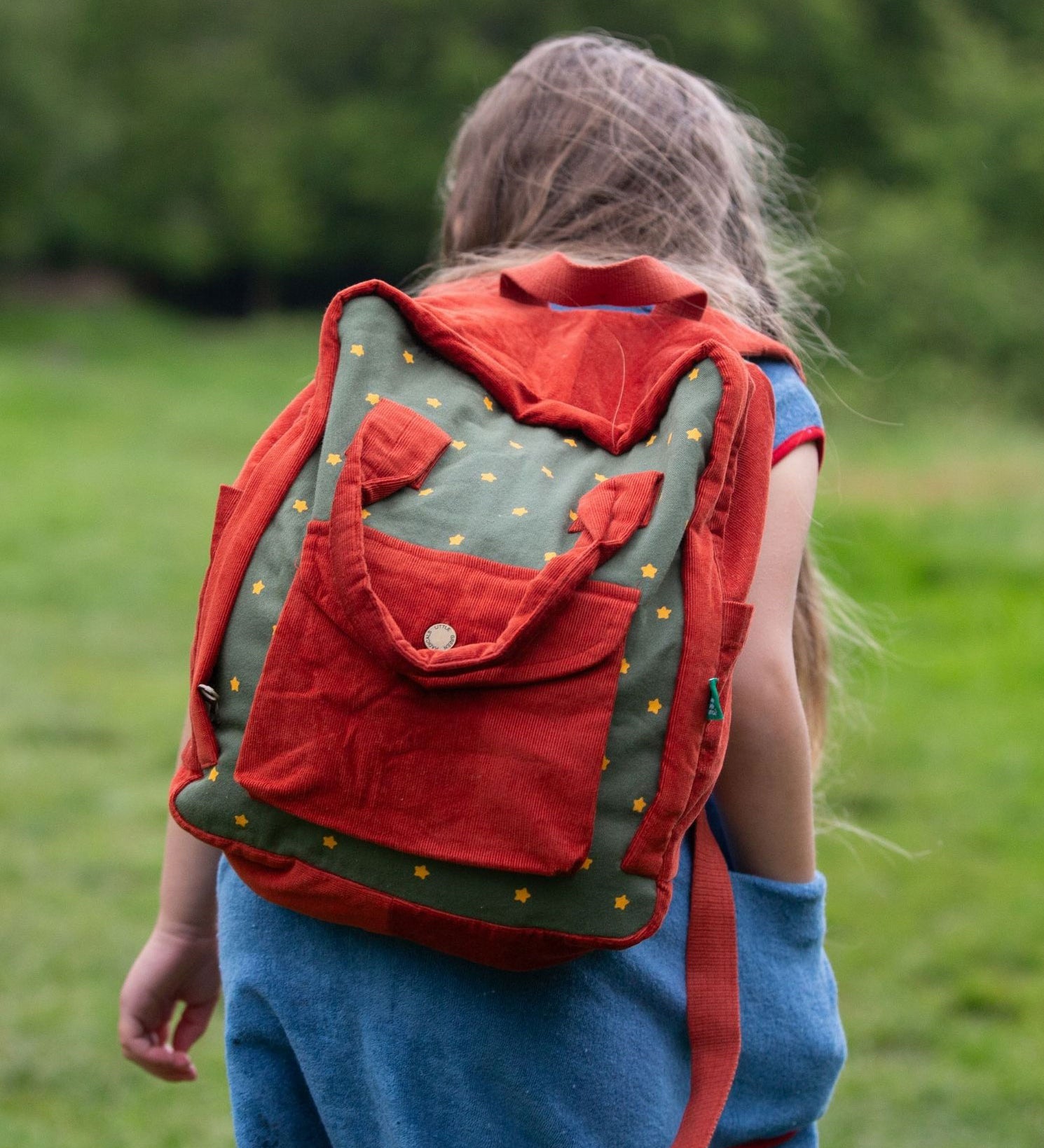 A child wearing the Little Green Radicals Kids Ready For Adventure Yellow Stars print red and green Backpack on their back. The child is facing away from the camera giving a full view of the backpack