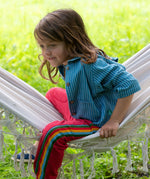 A child wearing the Little Green Radicals Red rainbow stripe Organic Cotton Comfy Joggers with a blue striped shirt. The child is sitting on a white hammock.