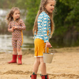 A child wearing the Little Green Radicals Children's Blue Whales design UPF 50+ Sunsafe Long Sleeve Rash Vest with yellow gold swim shorts and red wellingtons standing on a sandy beach.