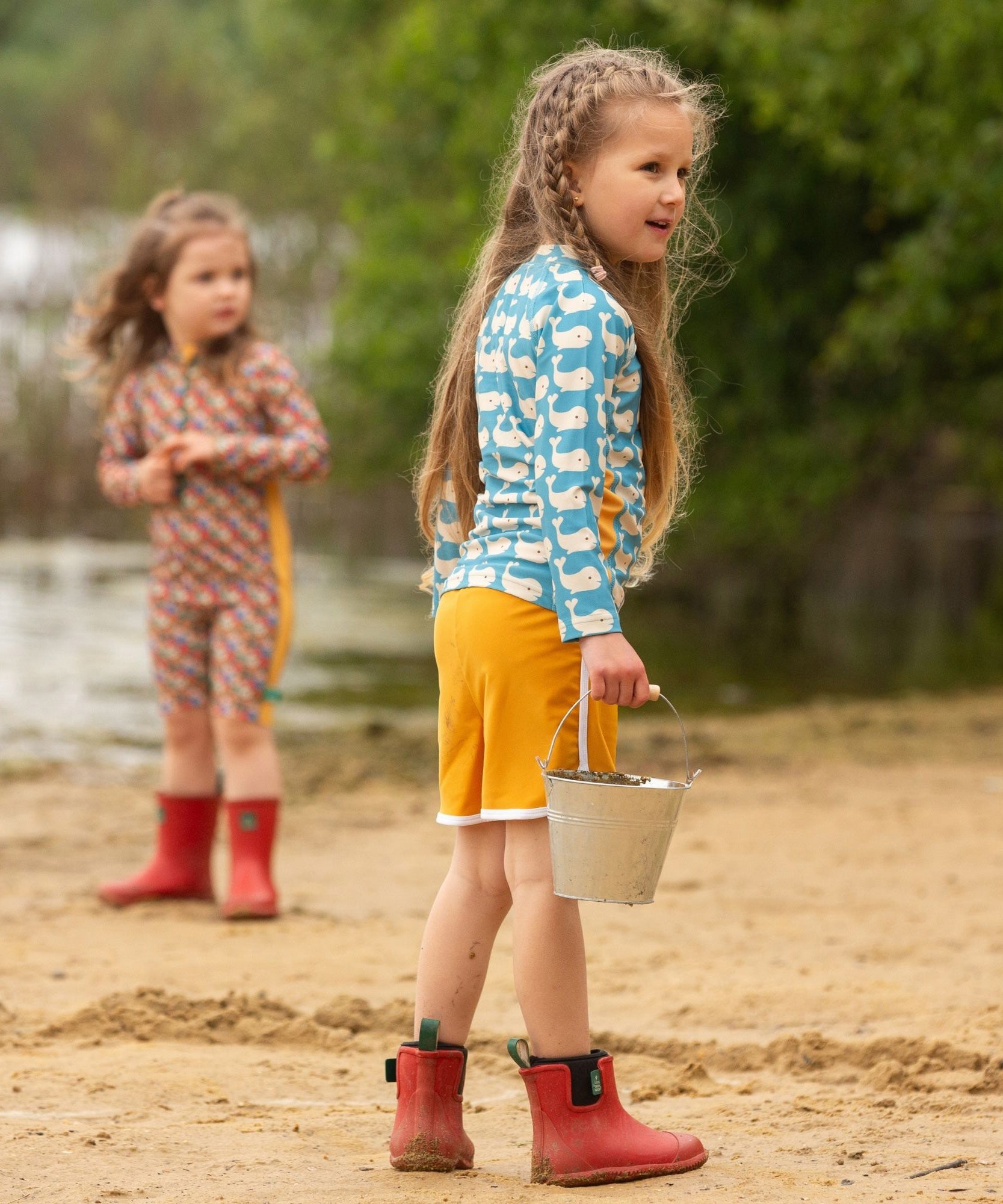 A child wearing the Little Green Radicals Children's Blue Whales design UPF 50+ Sunsafe Long Sleeve Rash Vest with yellow gold swim shorts and red wellingtons standing on a sandy beach.