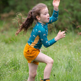 A child wearing the Little Green Radicals Teal Rainbows UPF 50+ Sunsafe Long Sleeve Rash Vest with yellow gold shorts and yellow wellingtons