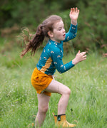 A child wearing the Little Green Radicals Teal Rainbows UPF 50+ Sunsafe Long Sleeve Rash Vest with yellow gold shorts and yellow wellingtons