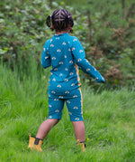 A child wearing the Little Green Radicals Teal Rainbows UPF 50+ Recycled Sunsafe Sunsuit with a pair of yellow gold wellingtons. The child is facing away from the camera showing the back of the suit.