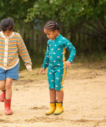 A child wearing the Little Green Radicals Teal Rainbows UPF 50+ Recycled Sunsafe Sunsuit with a pair of yellow gold wellingtons. The child is standing on sand at a beach. 
