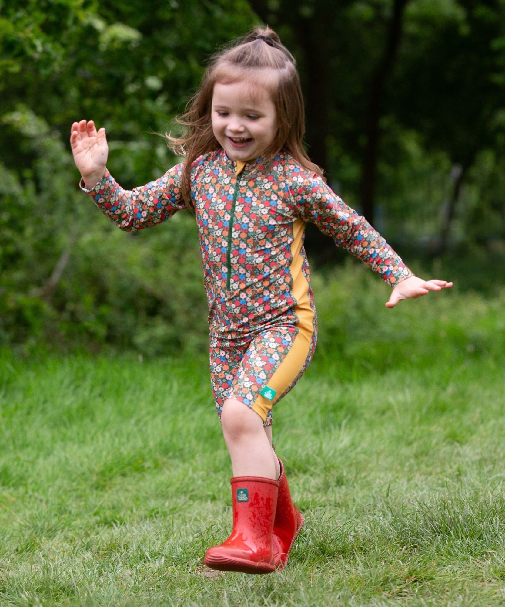 A child wearing the Little Green Radicals Vintage Flowers UPF 50+ Recycled Sunsafe Sunsuit with a pair of red wellington boots.