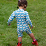 A child wearing the Little Green Radicals Blue Whales UPF 50+ Recycled Sunsafe Sunsuit with red wellington boots on grass outdoors. The child is facing away from the camera showing the back of the suit.