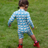 A child wearing the Little Green Radicals Blue Whales UPF 50+ Recycled Sunsafe Sunsuit with red wellington boots on grass outdoors. The child is facing away from the camera showing the back of the suit.