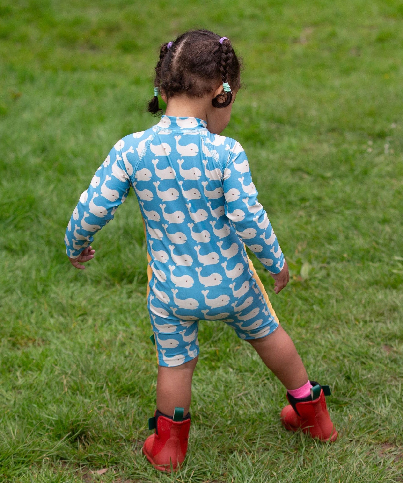 A child wearing the Little Green Radicals Blue Whales UPF 50+ Recycled Sunsafe Sunsuit with red wellington boots on grass outdoors. The child is facing away from the camera showing the back of the suit.