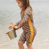 A child wearing the Little Green Radicals Vintage Flowers UPF 50+ Recycled Sunsafe Sunsuit with a pair of red wellington boots. They are standing in front of water with a bucket in their hands and their hands are covered in sand. 