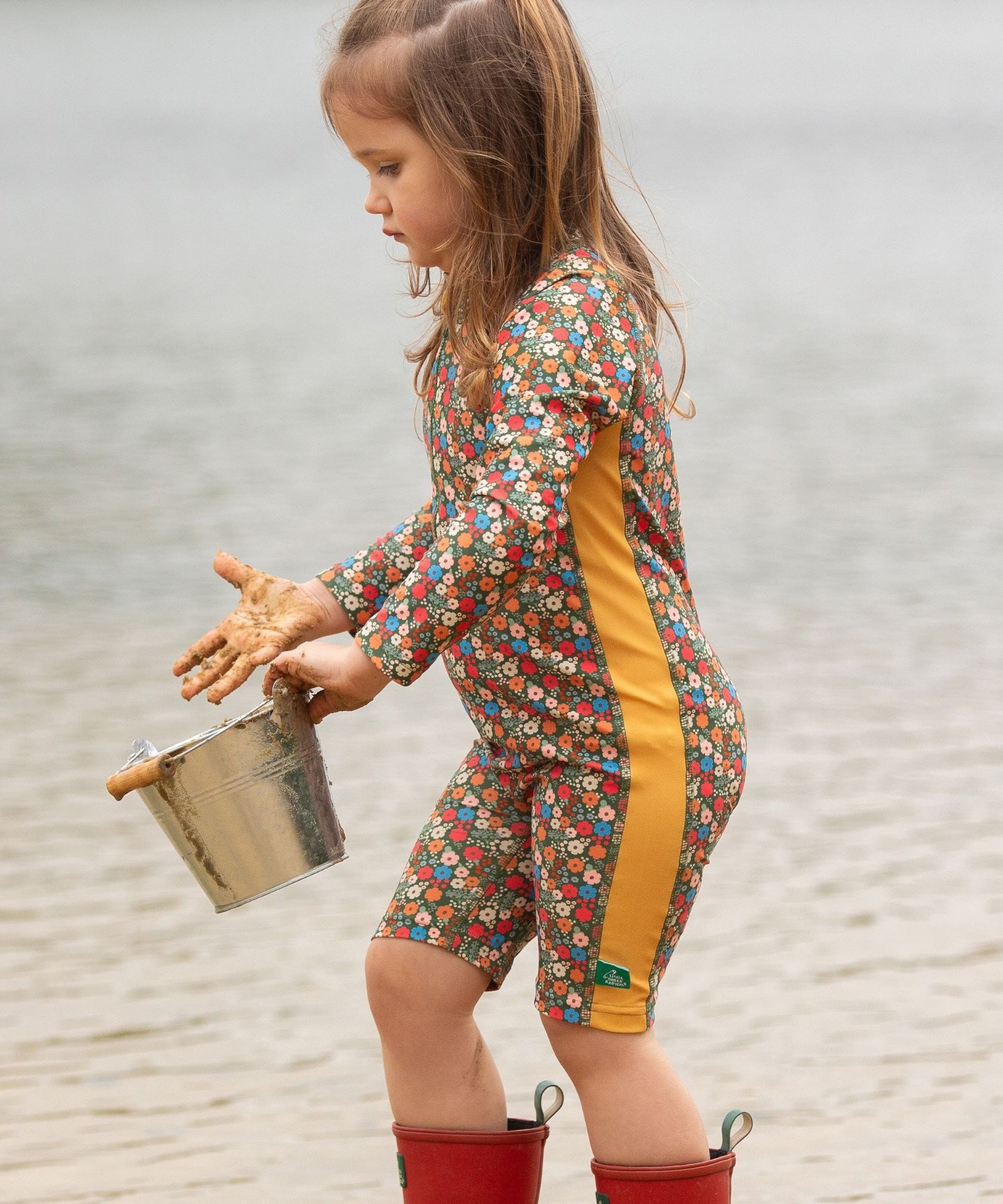 A child wearing the Little Green Radicals Vintage Flowers UPF 50+ Recycled Sunsafe Sunsuit with a pair of red wellington boots. They are standing in front of water with a bucket in their hands and their hands are covered in sand. 