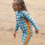 A child wearing the Little Green Radicals Blue Whales UPF 50+ Recycled Sunsafe Sunsuit on a sandy beach.
