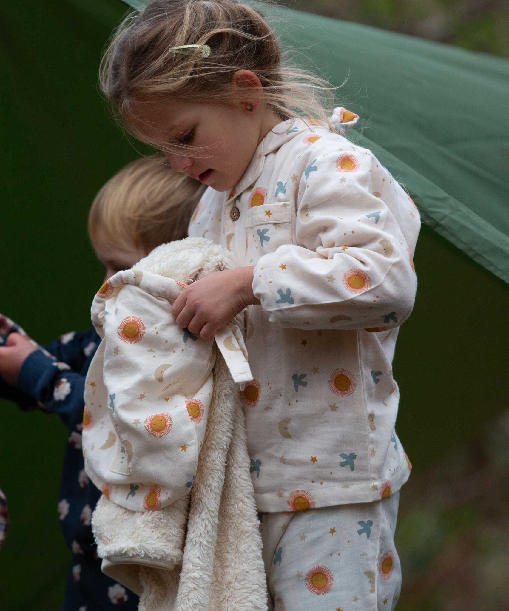A child wearing the Little Green Radicals moon and sun classic pyjama set holding a matching Sherpa lined hooded blanket. A GOTS organic cotton pyjama set including a long sleeve shirt style top in light pink with prints of cute birds flying through stars the moon and sun also includes matching bottoms. These pyjamas are part of a wide range of organic cotton clothing for children available here at Babipur.