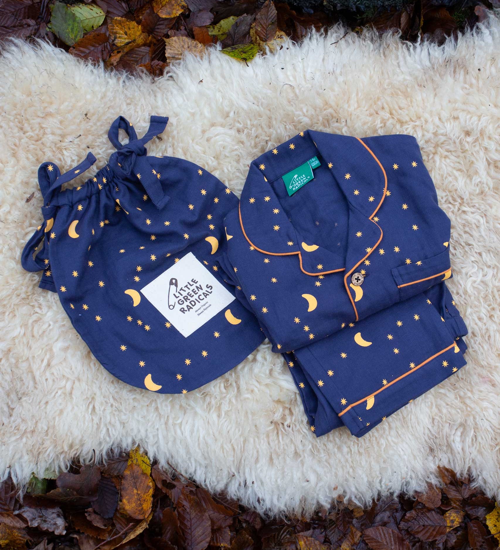 Little Green Radicals Dark Night  Organic Cotton Classic children's Pyjamas placed folded on a sheepskin rug. These navy blue traditional style pyjamas have a yellow moon and star print all over. These pyjamas are part of a wide range of organic cotton clothing for children available here at Babipur.