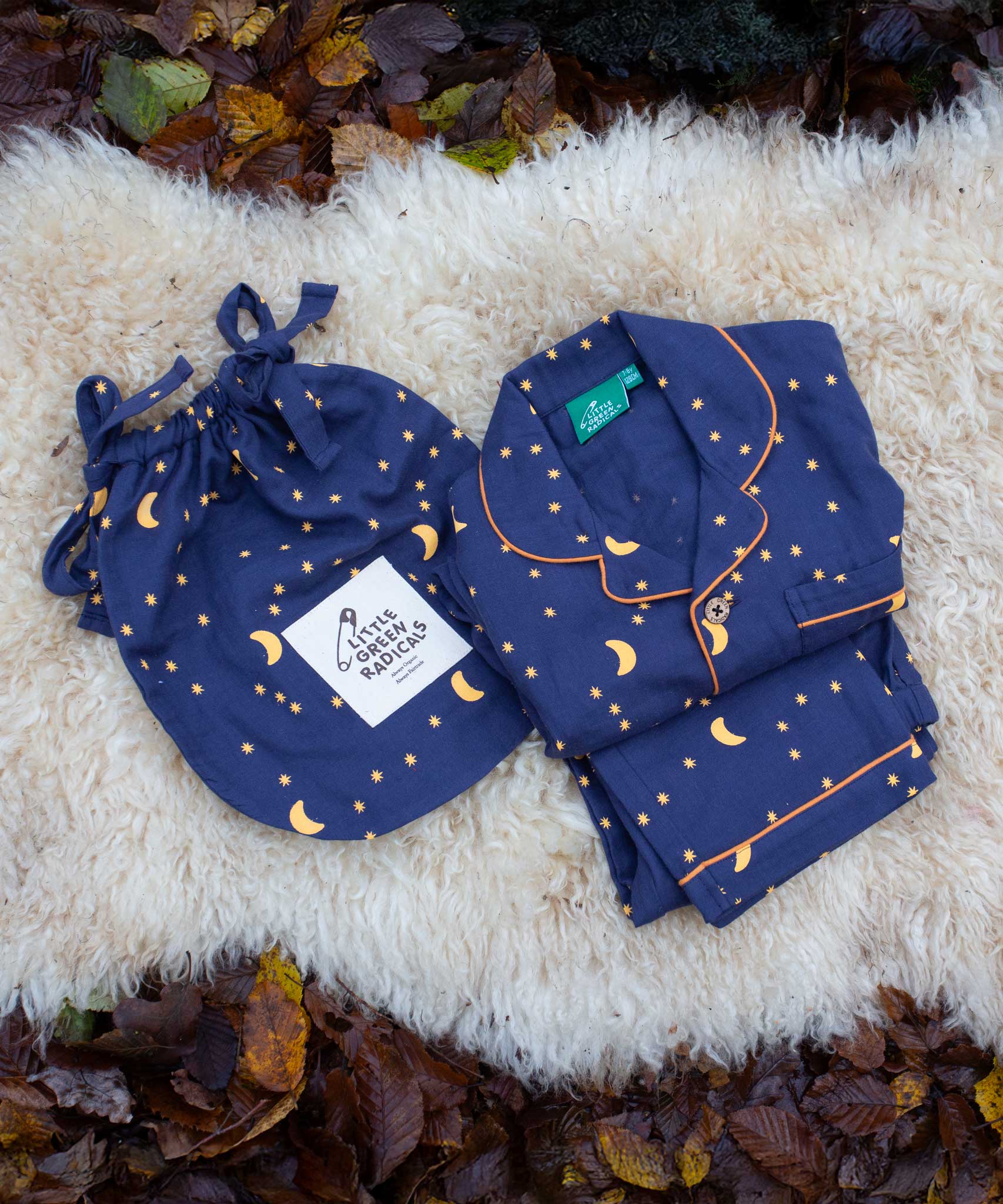 Little Green Radicals Dark Night  Organic Cotton Classic children's Pyjamas placed folded on a sheepskin rug. These navy blue traditional style pyjamas have a yellow moon and star print all over. These pyjamas are part of a wide range of organic cotton clothing for children available here at Babipur.