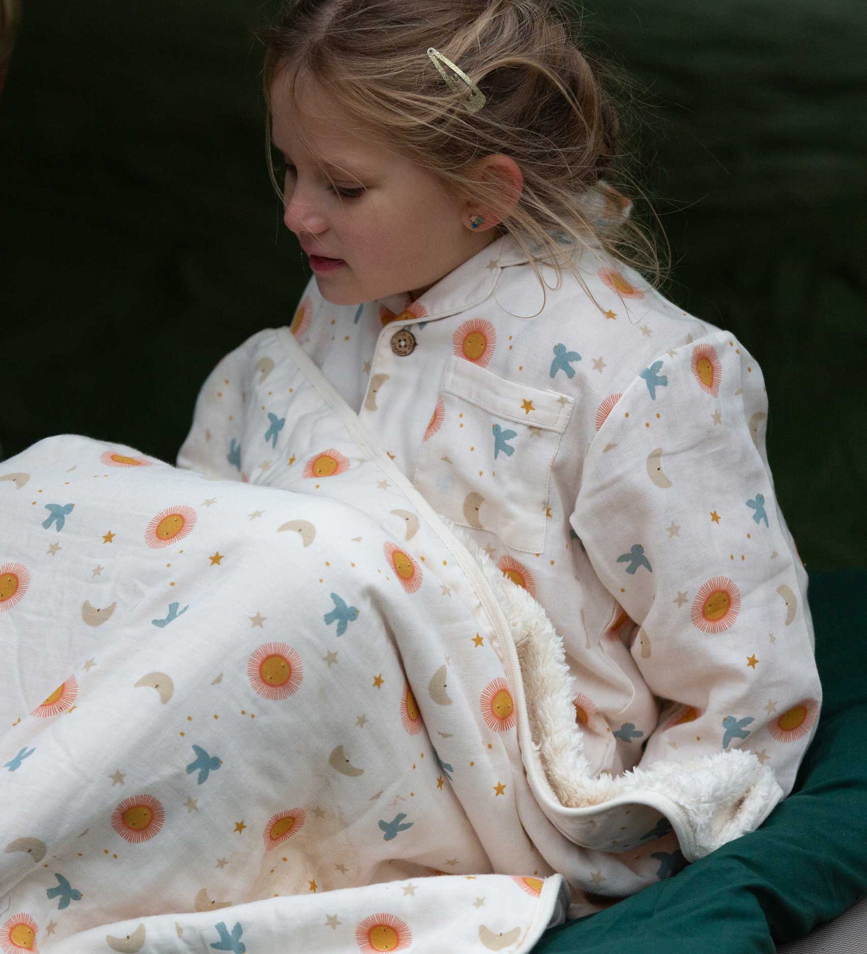 A child wearing the Little Green Radicals moon and sun classic pyjama set. A GOTS organic cotton pyjama set including a long sleeve shirt style top in light pink with prints of cute birds flying through stars the moon and sun also includes matching bottoms. These pyjamas are part of a wide range of organic cotton clothing for children available here at Babipur.