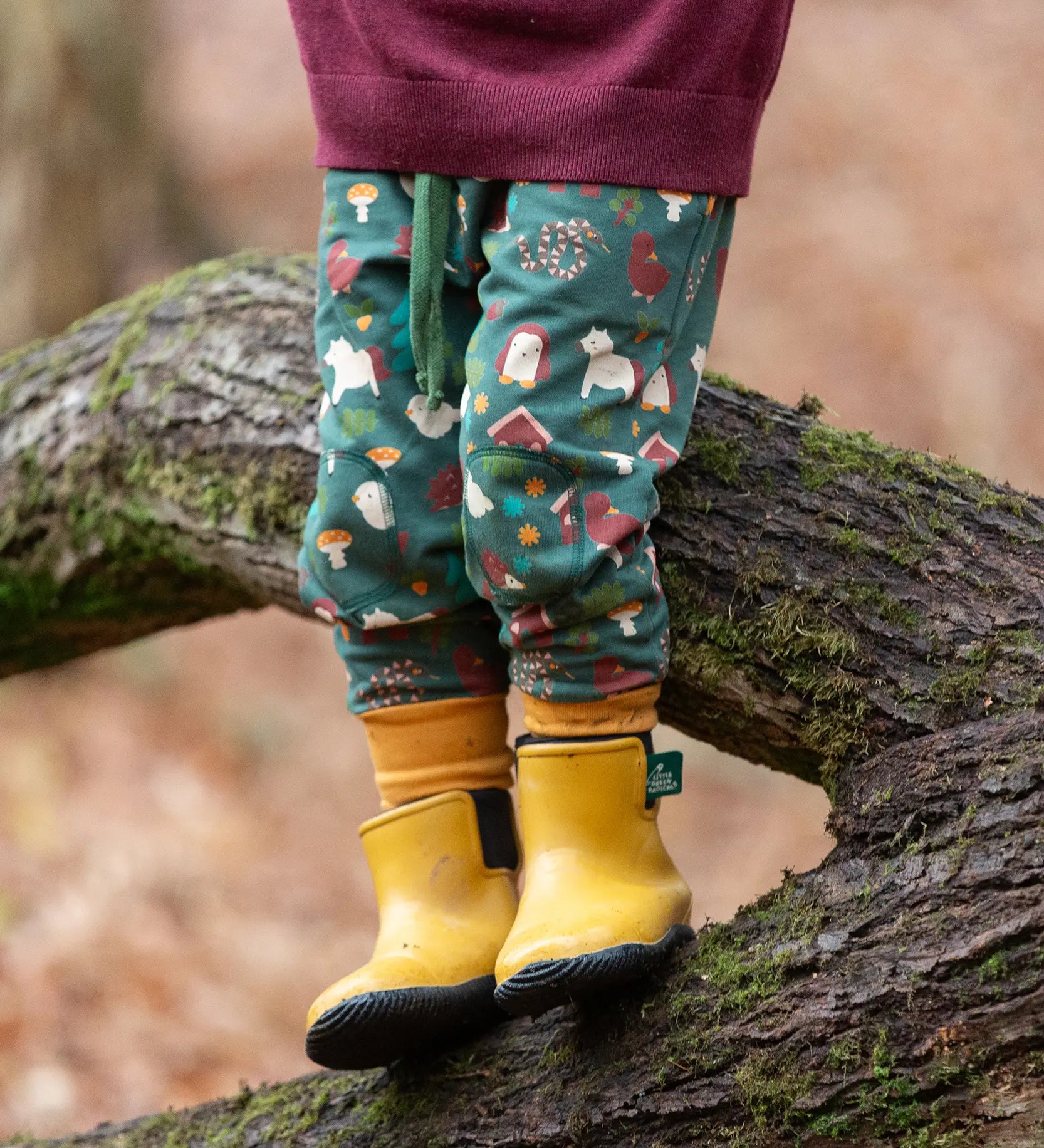 Child wearing the Little Green Radicals cosy organic cotton knee patch joggers with a woodland design print. Dark green joggers with a woodland print featuring animals, trees a hut.