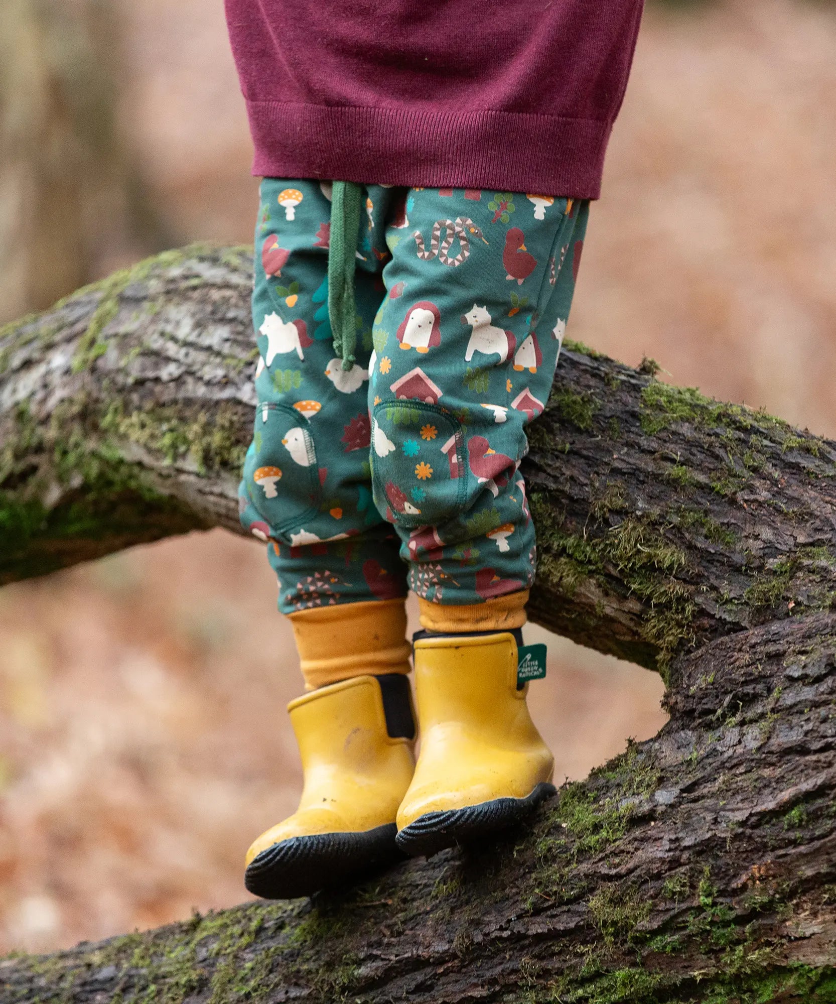 Child wearing the Little Green Radicals cosy organic cotton knee patch joggers with a woodland design print. Dark green joggers with a woodland print featuring animals, trees a hut.